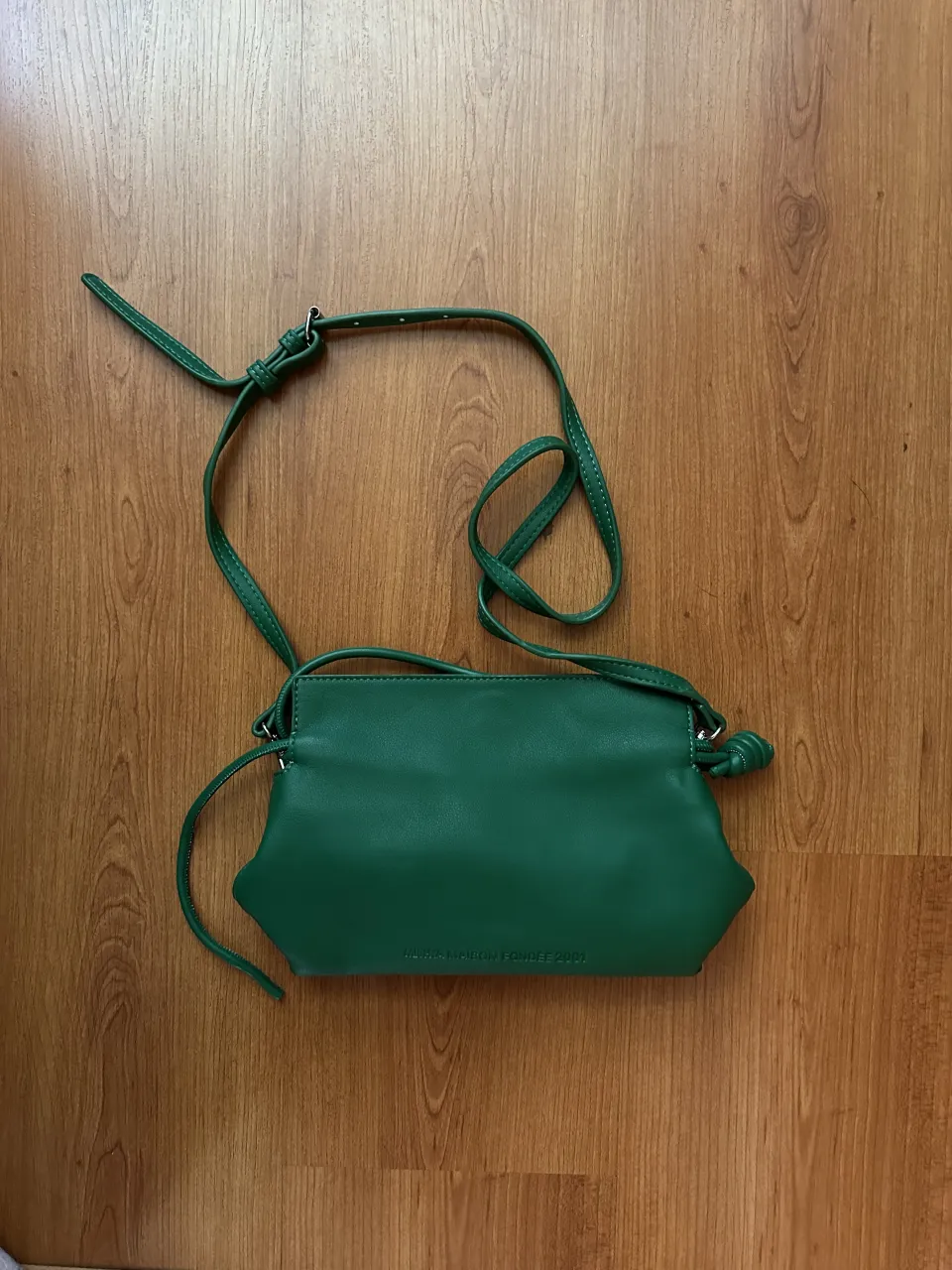 Cartera verde con correa larga ajustable y detalle de nudo en uno de los extremos. Posee inscripción en relieve en la parte frontal.