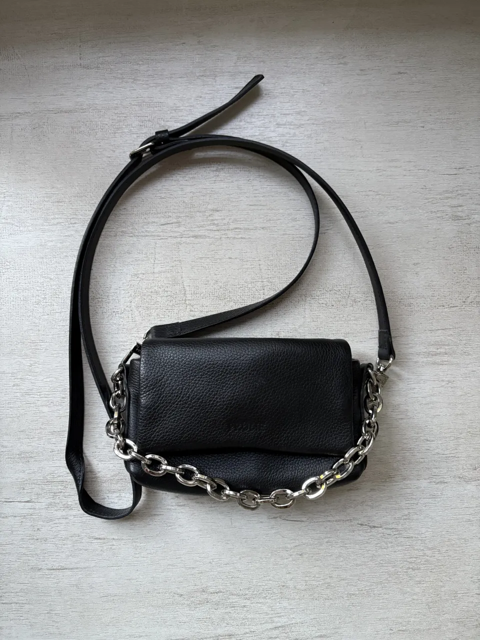 Cartera negra de cuero con cadena metálica plateada. Tiene correa larga ajustable y cierre de botón a presión.