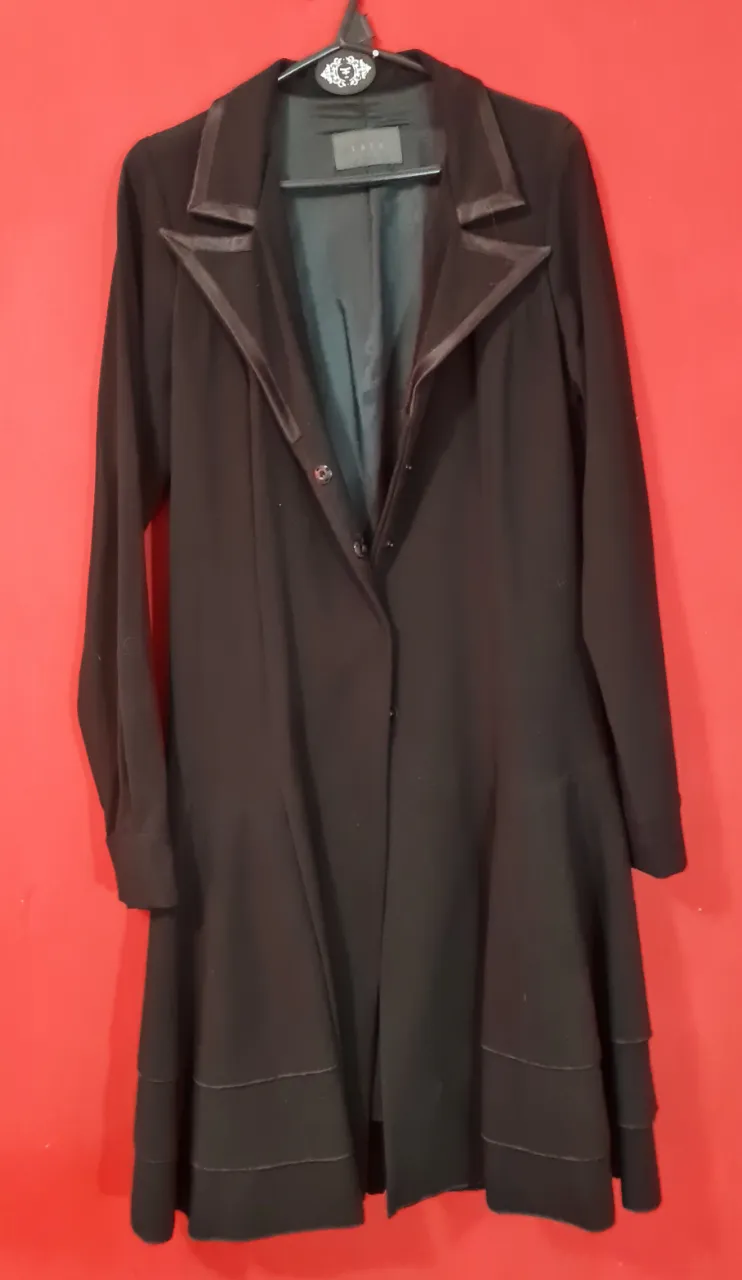 Tapado negro largo con solapas y detalles de satén. Posee botones al frente y un corte acampanado con volados en la parte inferior. Ideal para un look elegante y sofisticado.