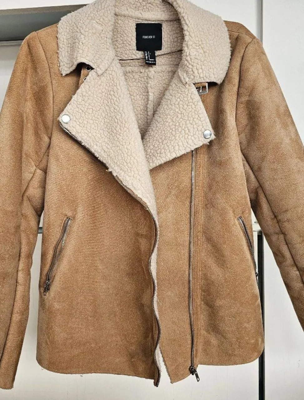 Campera de gamuza sintética color marrón con cuello y forro de corderito color crudo. Cierre metálico al frente y bolsillos laterales con cierre. Puños con detalle de cierre.