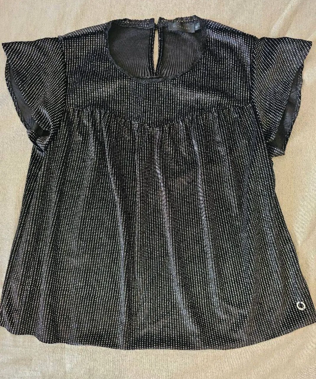 Remera negra con detalles plateados brillantes en forma de finas líneas verticales. Posee mangas cortas con volados y cuello redondo.