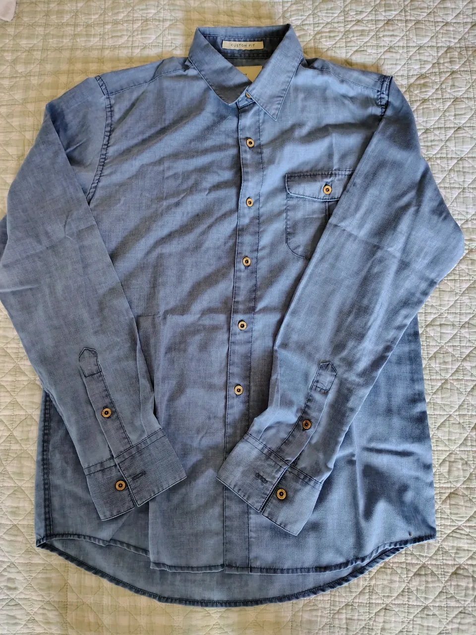 Camisa de jean de manga larga, color azul. Posee botones dorados en el frente y en los puños. Tiene un bolsillo en el pecho.