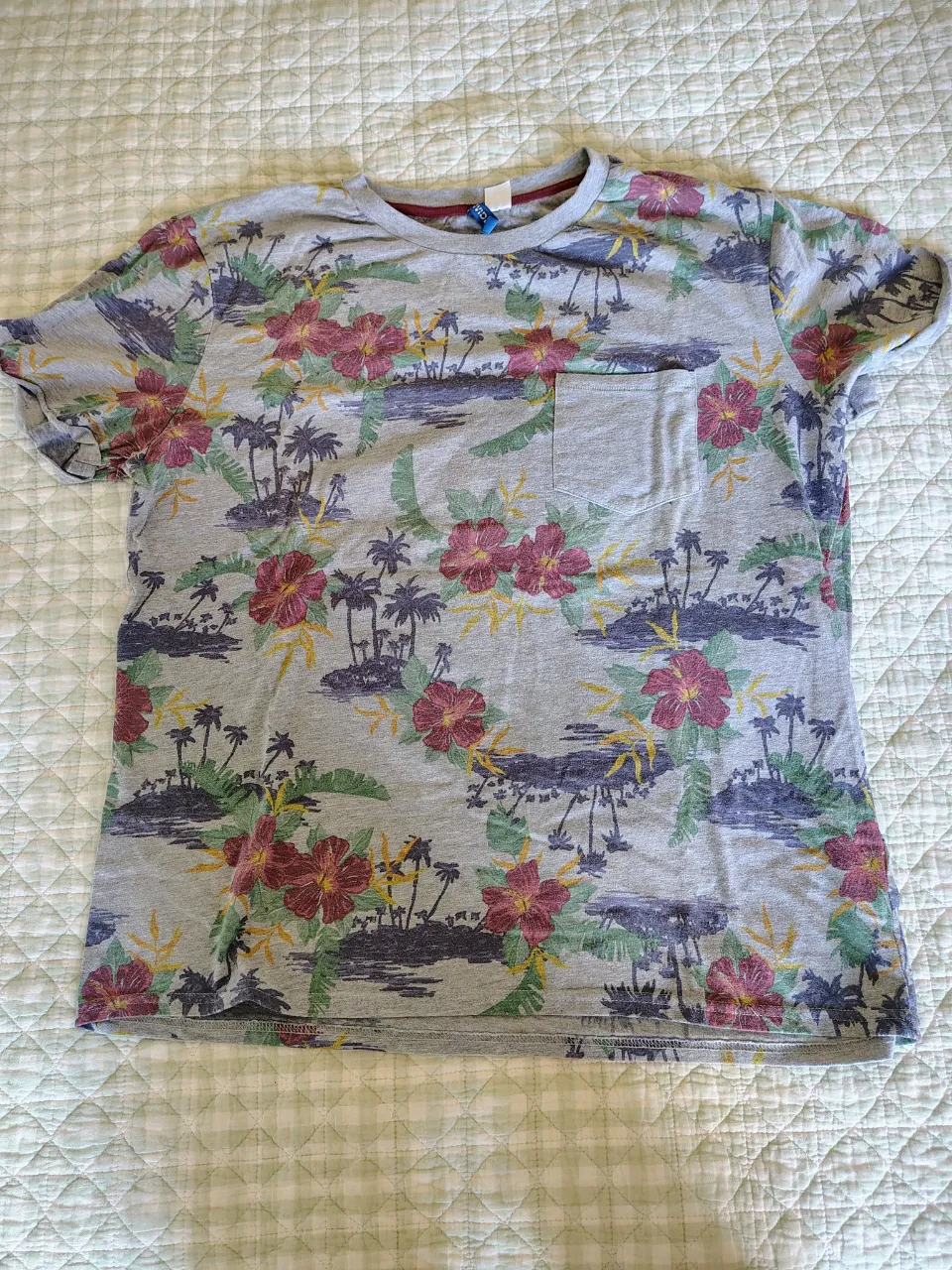 Remera gris con estampa de palmeras, flores y paisaje tropical. Tiene un bolsillo en el frente.