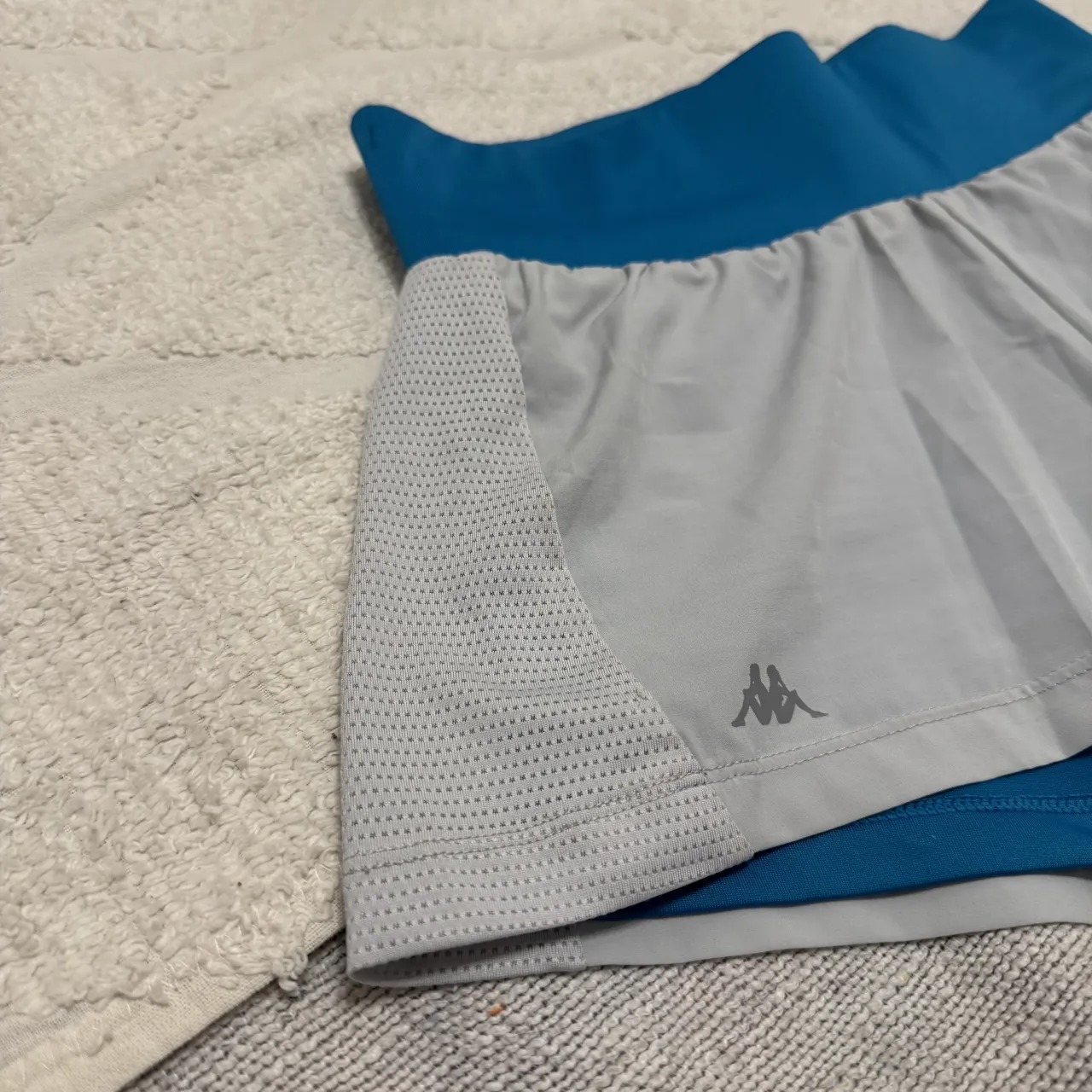 Shorts deportivos de color azul y gris. Tienen un bolsillo con cierre en la parte trasera y un logo de la marca Kappa en la parte delantera y trasera.