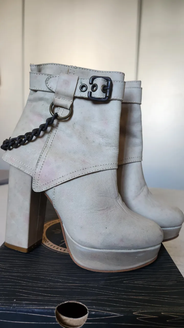 Botas de cuero color crudo con plataforma y taco alto. Tienen una hebilla y una cadena decorativa en la parte superior.