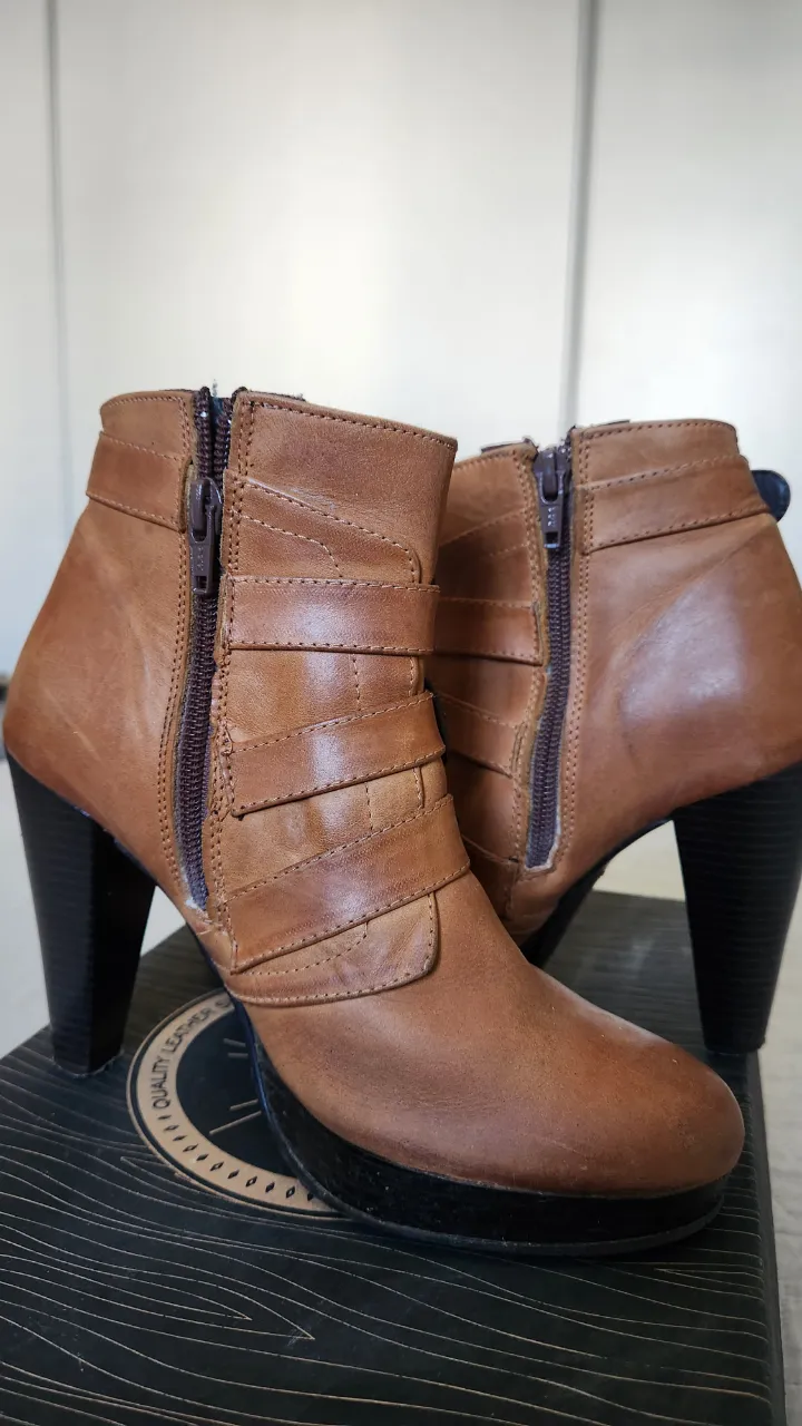 Botitas de cuero color marrón con taco alto y plataforma. Tienen cierre lateral y tres hebillas decorativas con herrajes metálicos. La suela es de goma.