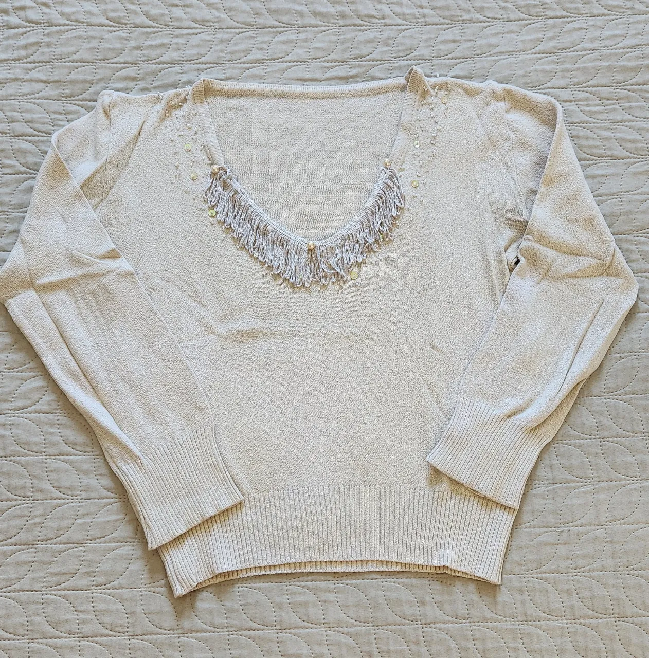 Sweater de hilo color crudo con escote en V adornado con flecos y mostacillas. Posee detalles de mostacillas y lentejuelas en la zona del escote y hombros.