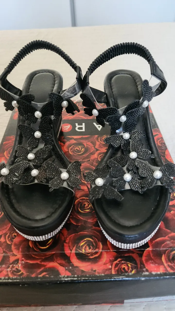 Sandalias negras con plataforma y taco cuña. Tienen tiras finas transparentes y apliques de mariposas y flores negras con perlas blancas. La base de la plataforma y el taco están decorados con strass plateado.