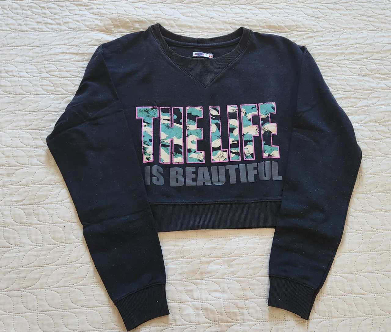 Buzo negro crop con estampa "THE LIFE IS BEAUTIFUL" en el frente. La estampa "THE LIFE" tiene letras con un diseño camuflado en tonos verdes, rosas y beige, y "IS BEAUTIFUL" está en letras grises.