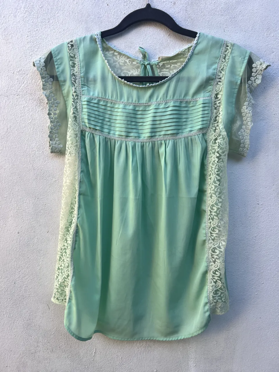 Remera color verde menta con detalles de encaje blanco en los laterales y mangas. Posee cuello redondo y lazo en la espalda para atar.