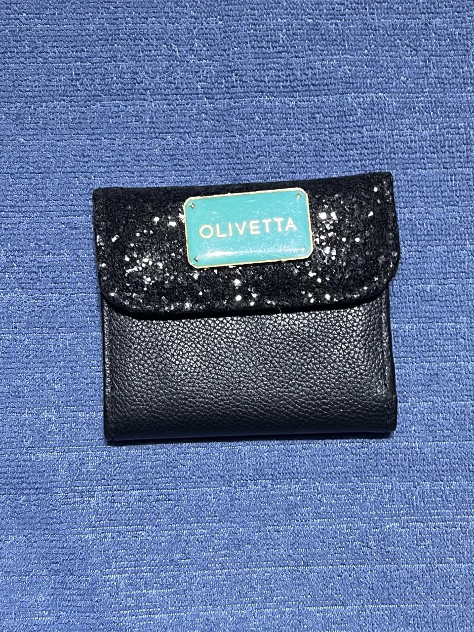 Billetera negra con tapa de brillos  y detalle de placa rectangular turquesa con la inscripción "OLIVETTA". Al abrirla, se observan múltiples compartimentos para tarjetas.