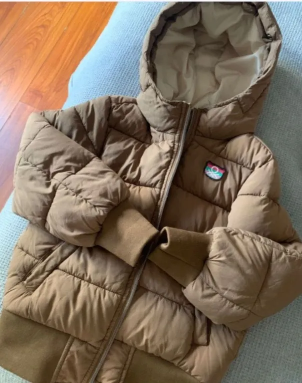 Campera acolchada marrón con capucha. Tiene cierre al frente y un parche con un logo circular en el pecho izquierdo. Los puños y la parte inferior de la campera son de ribb.