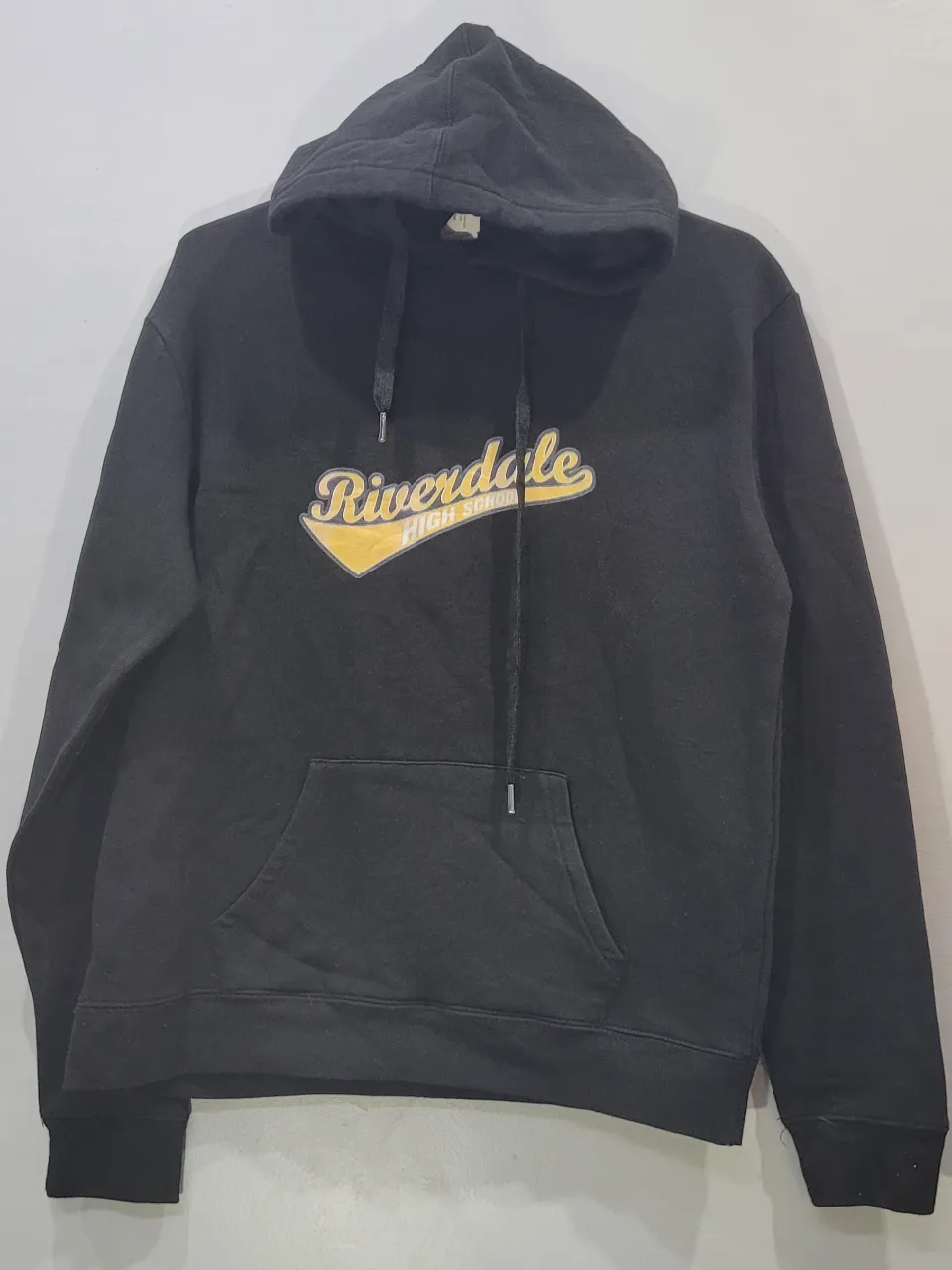 Buzo negro con capucha y cordones. Estampado frontal con texto "Riverdale High School" en amarillo y blanco.