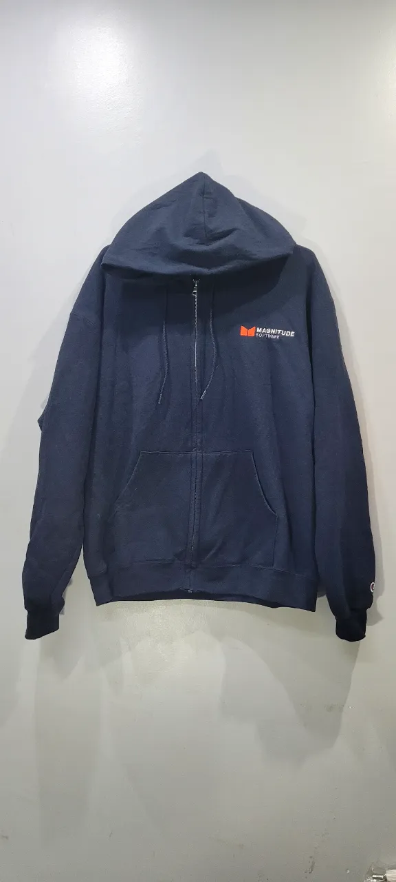Buzo azul marino con capucha y cierre completo. Tiene bolsillos frontales tipo canguro y un logo naranja y blanco en el frente izquierdo. En la manga izquierda tiene un pequeño logo bordado de la marca Champion.