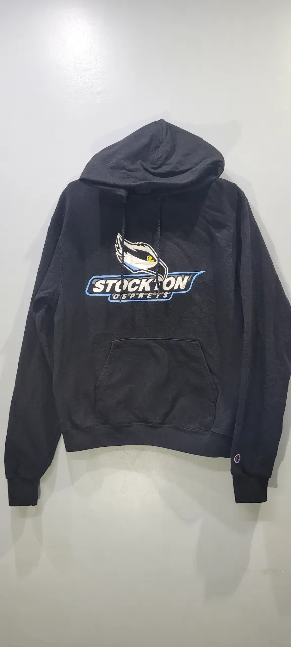 Buzo negro con capucha y bolsillo canguro. Estampado frontal con el logo de "Stockton Ospreys". Logo de Champion en la manga.