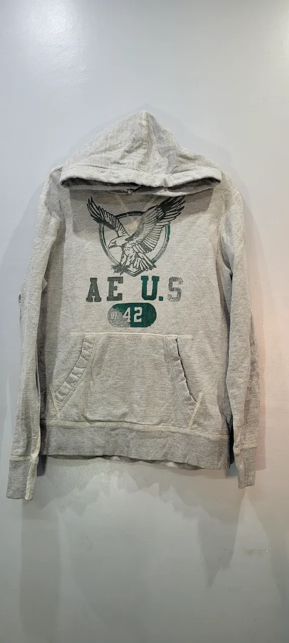 Buzo gris con capucha y estampa de águila y texto "AE U.S 42" en el frente. Tiene bolsillo canguro.
