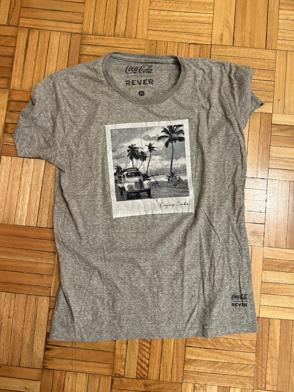 Remera de hombre gris con estampado de Coca-Cola. Diseño retro con imagen de camioneta en la playa y la frase 'Enjoy Coke'. Cuello redondo y manga corta. Ideal para un look casual y con onda. Un solo uso.