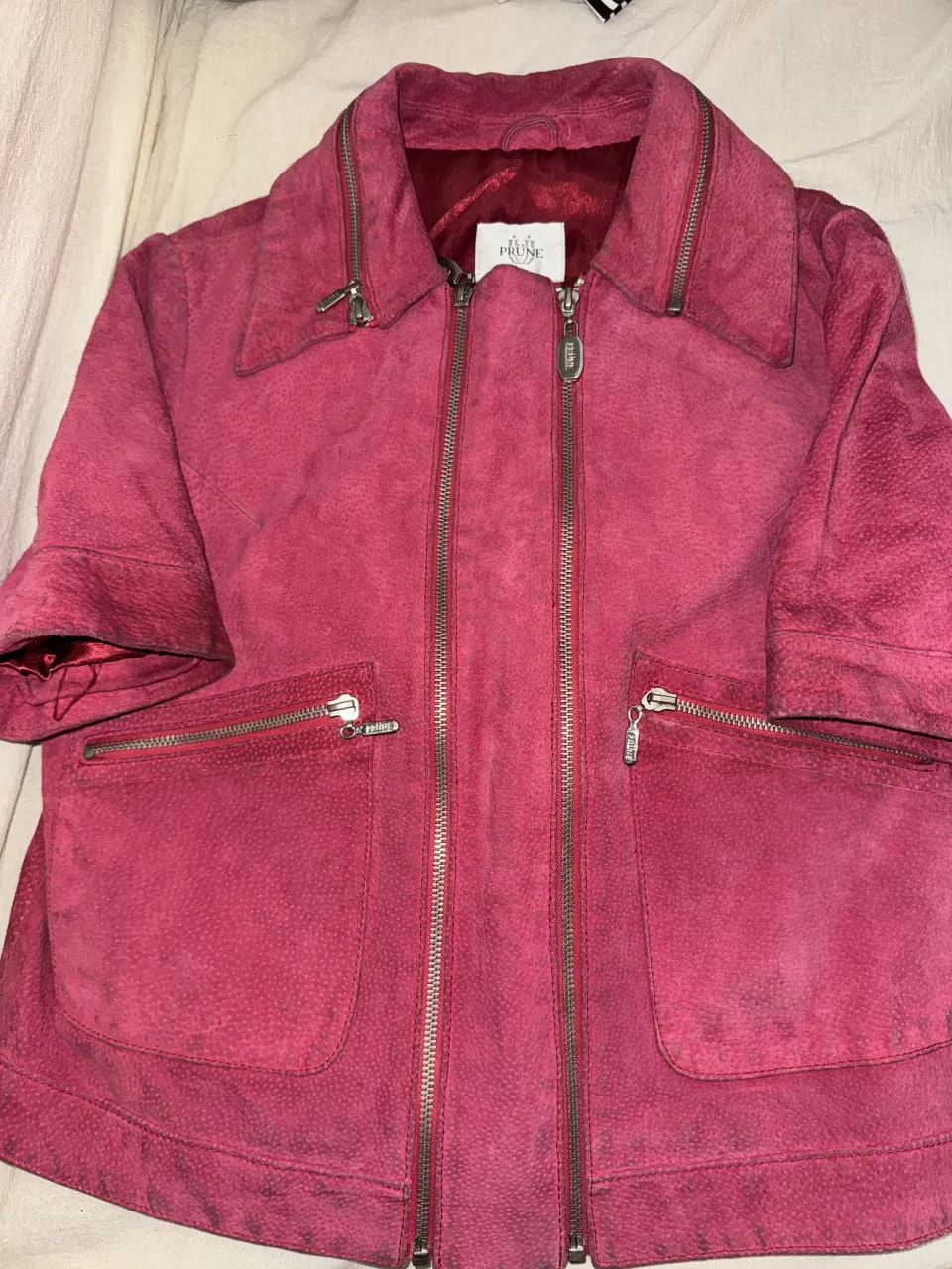 Campera corta de cuero de gamuza marca Prune color fucsia con cuello solapa. Posee doble cierre central y dos bolsillos frontales con cierre. Los puños son cortos y la espalda tiene un corte central con dos botones.