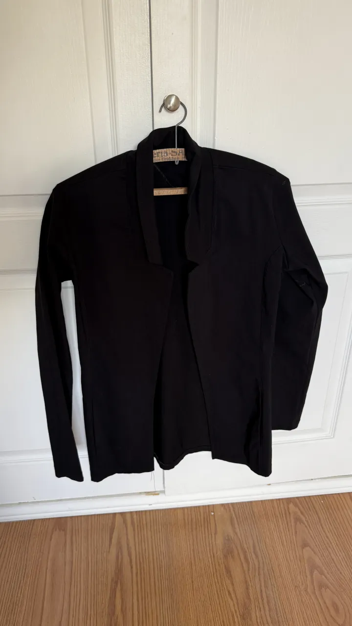 Blazer negro de manga larga con cuello solapa y corte abierto al frente. Presenta costuras visibles en la parte delantera y en las mangas.