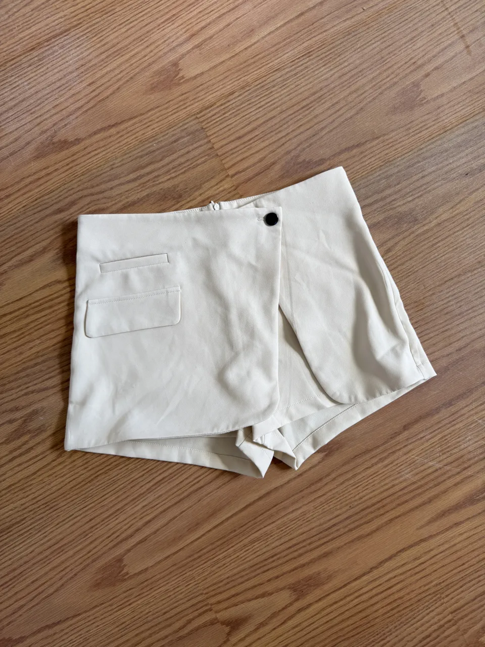 Shorts color blanco roto con detalle de un botón negro en la parte delantera y un bolsillo falso con pespuntes.