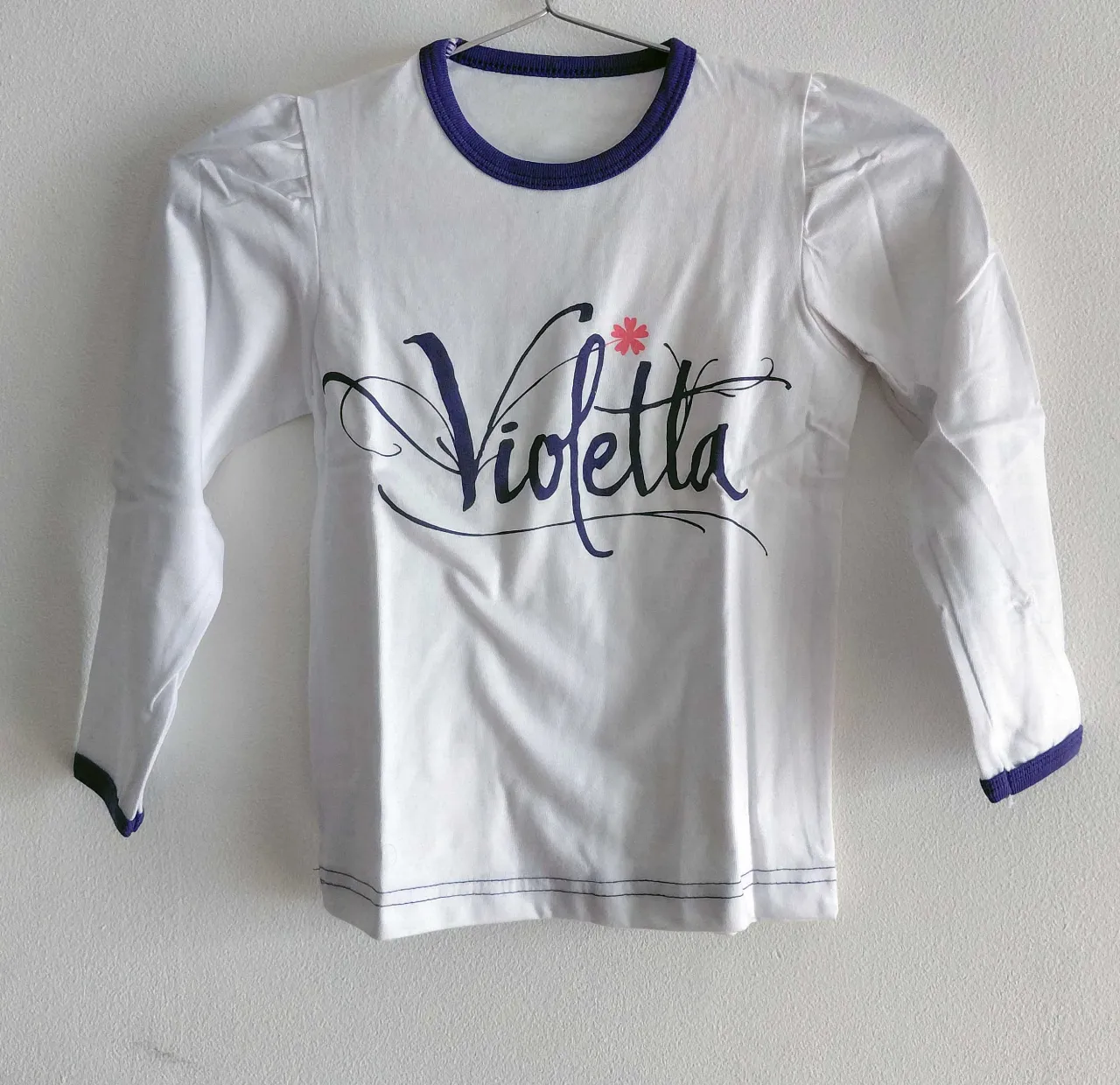 Remera blanca de manga larga con cuello redondo y ribete azul. Estampa frontal con la palabra "Violetta" en letras cursivas azules y una pequeña flor rosa. Puños y ruedo con detalles de costura azul. En la espalda, un parche cuadrado con un logo circular que incluye un globo terráqueo y la inscripción "planeta escabeche 5-6 AÑOS HECHO ACA".