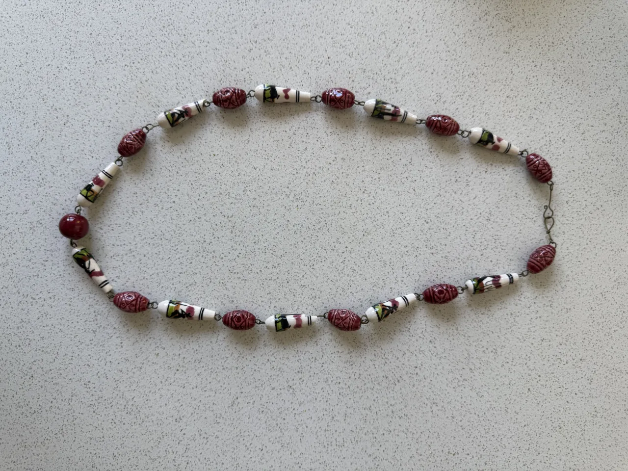 Collar delicado con cuentas de cerámica pintadas a mano. Presenta un diseño único con motivos florales y geométricos en tonos borgoña y blanco, con un detalle de cuenta roja vibrante. Ideal para darle un toque especial a cualquier outfit.