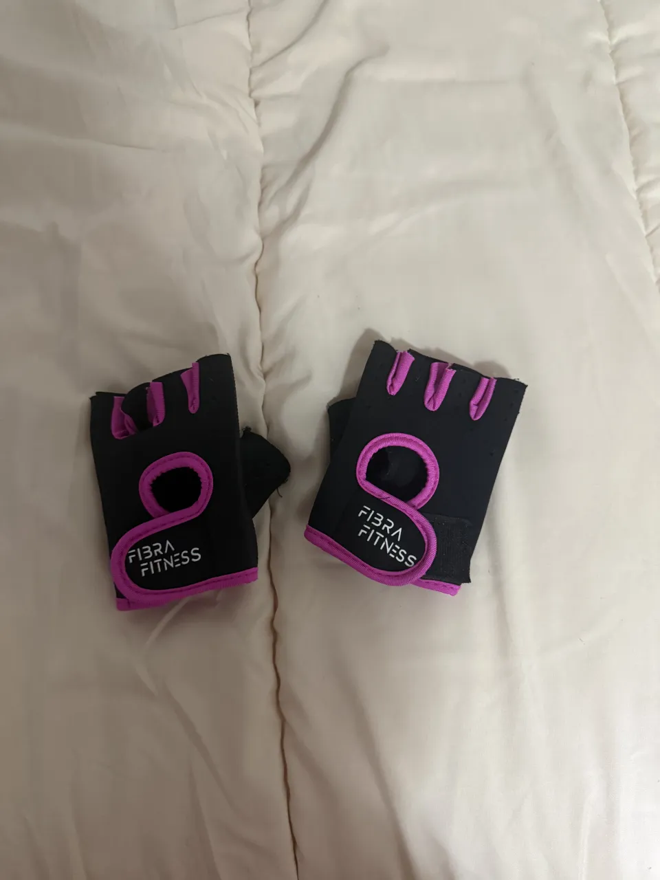 Par de guantes de entrenamiento sin dedos, color negro con detalles en fucsia. Tienen la inscripción "FIBRA FITNESS" en blanco.