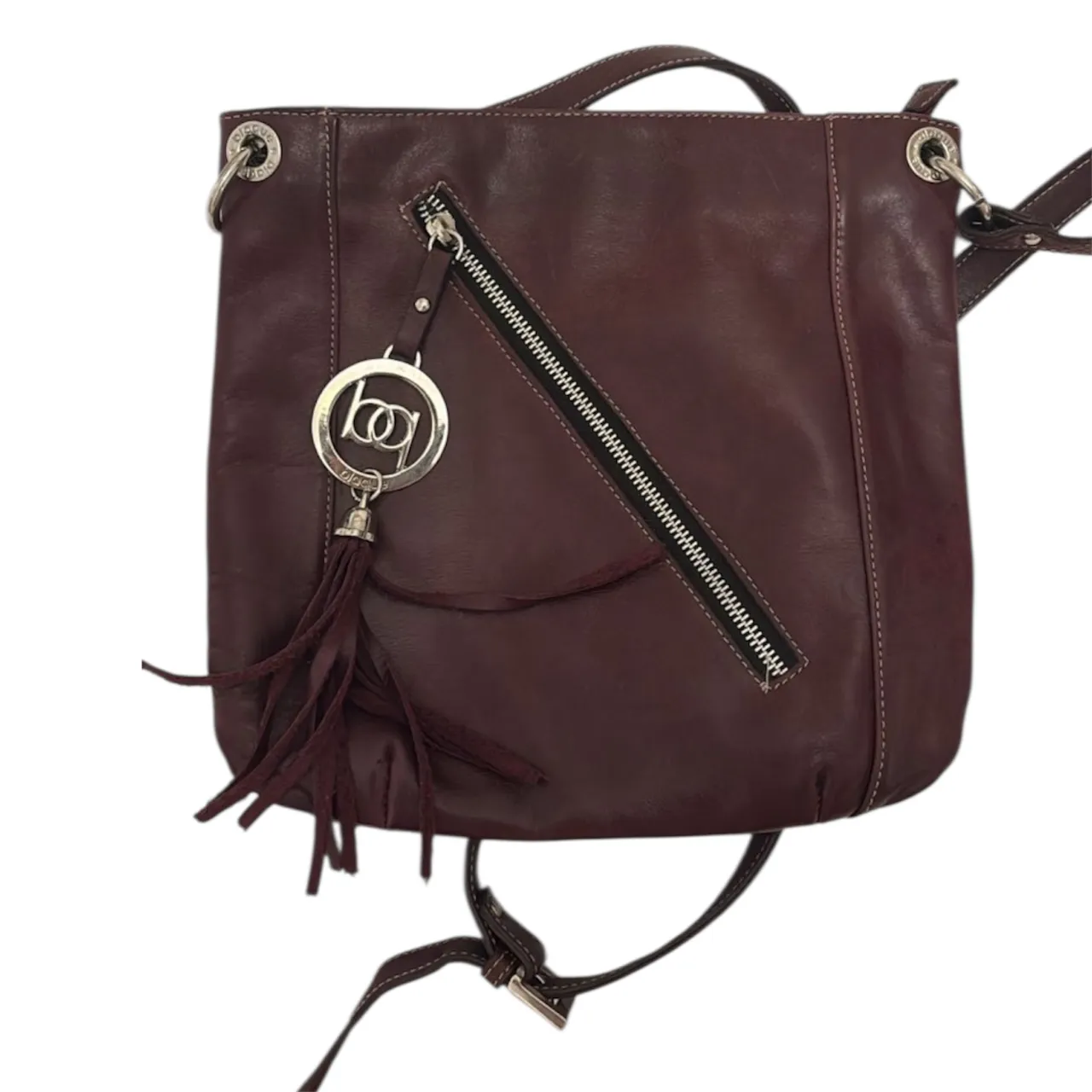 Bolso de cuero color bordó con detalle de costuras blancas. Posee un bolsillo exterior con cierre diagonal y un dije con flecos. El interior está forrado con un estampado de hojas.