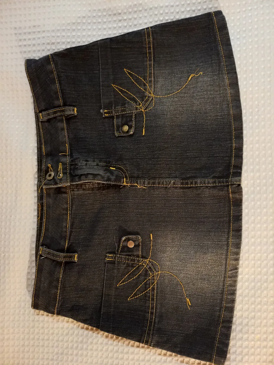 Pollera de jean corta, color negro con pespuntes amarillos. Tiene bolsillos delanteros y traseros con detalles bordados.