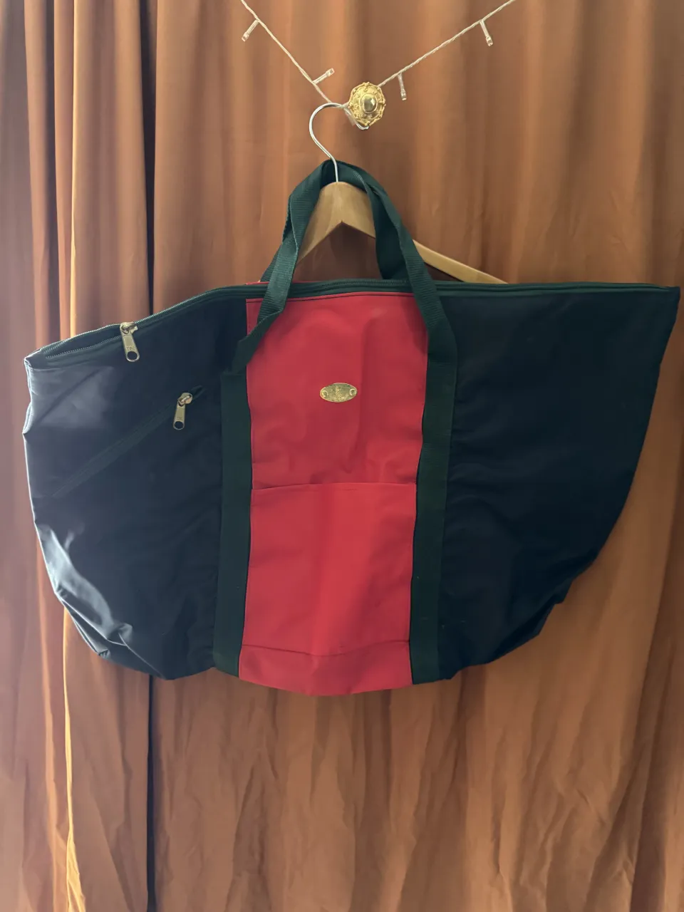 Bolsa de viaje grande, con dos asas cortas y un bolsillo exterior con cierre diagonal. Presenta un panel frontal rojo con un bolsillo abierto y un logo ovalado dorado. El interior es de color rojo y negro.