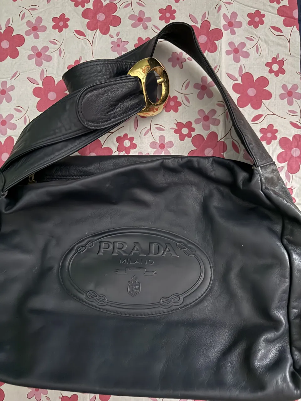 Bolso negro de cuero con logo PRADA MILANO grabado en el frente. Correa con hebilla dorada. Interior forrado con tela estampada con el logo de la marca.