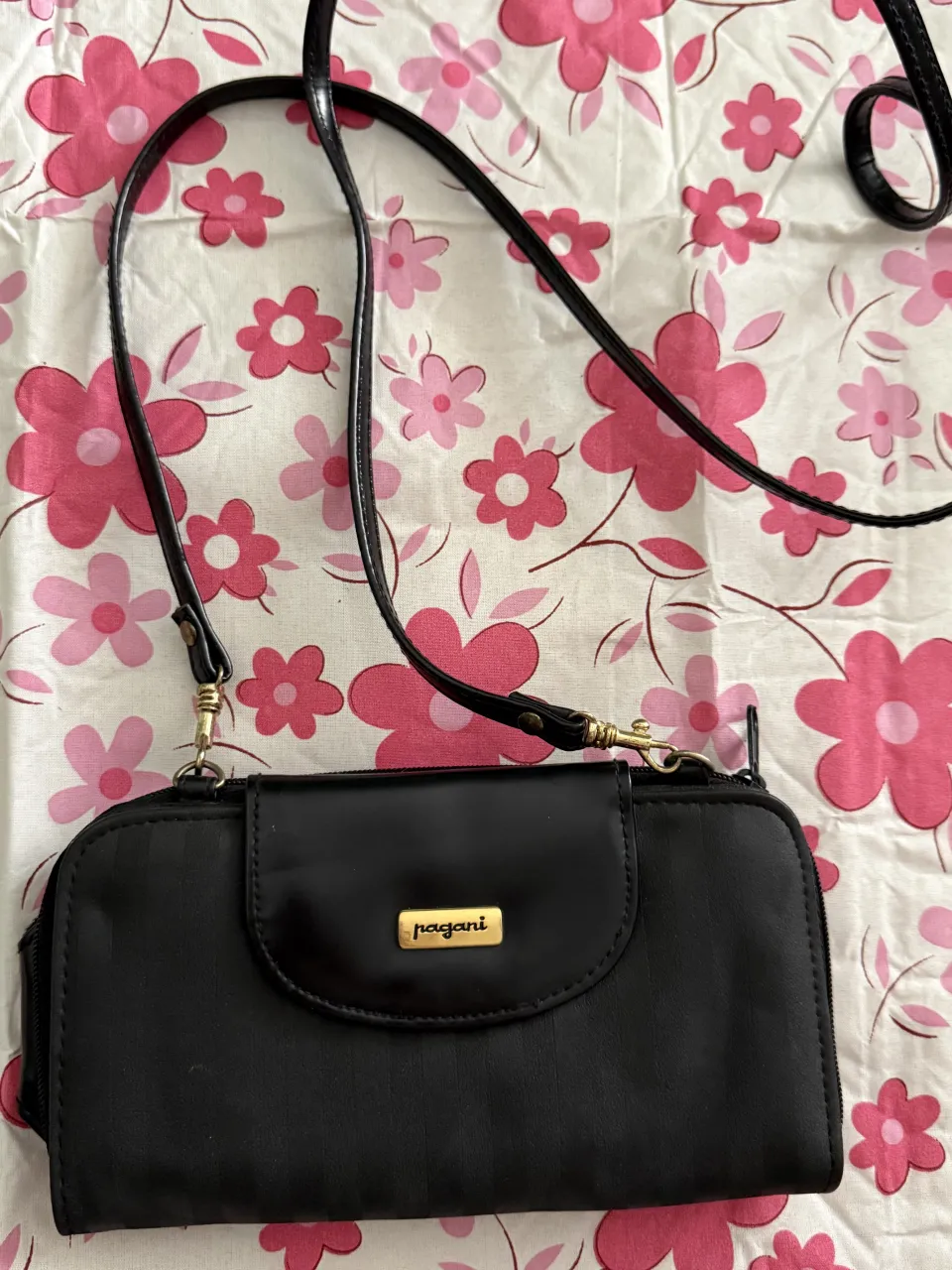 Cartera negra con correa larga y detalles de costuras finas. Posee un bolsillo frontal con solapa y logo dorado de la marca 'pagani'. Se abre mediante cierre y tiene múltiples compartimentos internos para tarjetas y documentos.