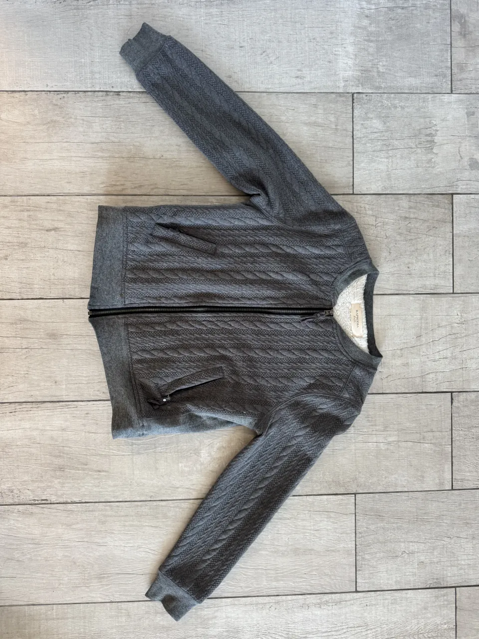 Buzo gris con cierre completo y bolsillos delanteros. Posee un tejido texturizado con diseño de trenzas.