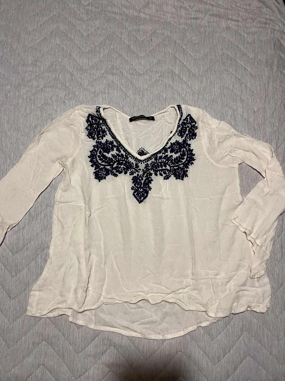 Blusa blanca con mangas largas y escote en V. Presenta un bordado floral en color azul marino en la zona del escote y la parte superior del pecho. El tejido es ligero y con textura.