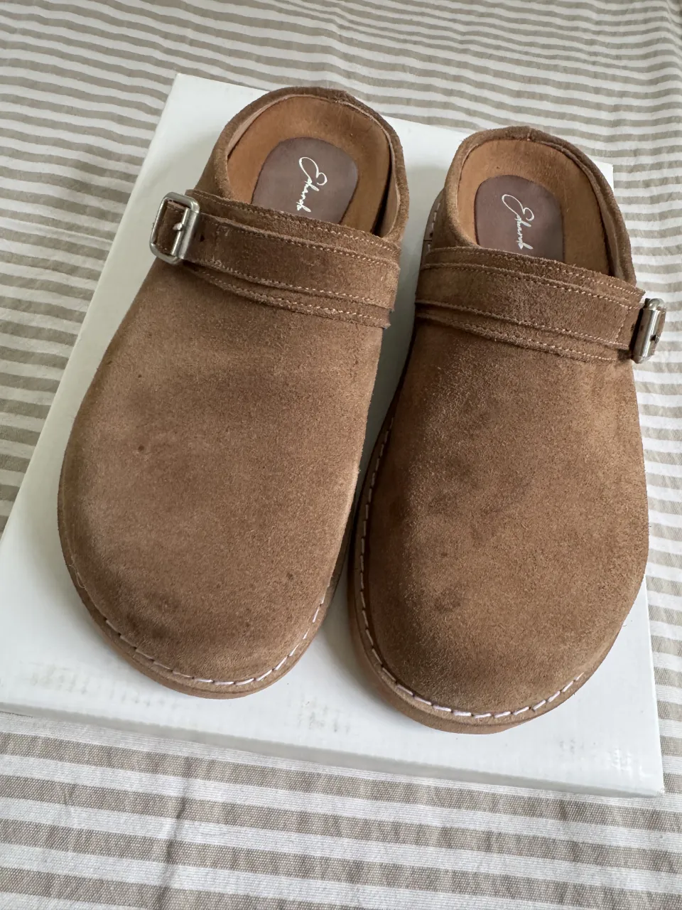 Zuecos de gamuza color marrón. Tienen una hebilla metálica en la tira superior y costuras blancas al tono en el borde. Precio original 150.000 solo tienen un uso 