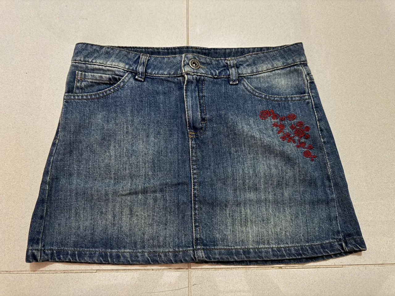 Pollera de jean azul con bordado de flores rojas en la parte delantera y trasera. Tiene bolsillos.