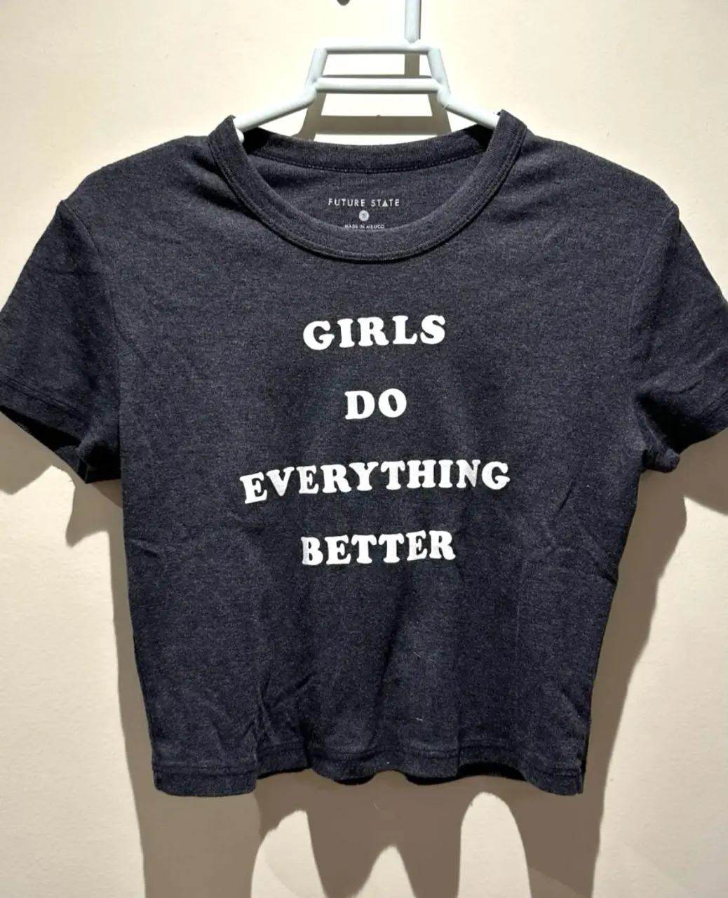 Remera de mujer de color gris oscuro con la frase "GIRLS DO EVERYTHING BETTER" en letras blancas. Corte crop, ideal para un look casual y moderno.