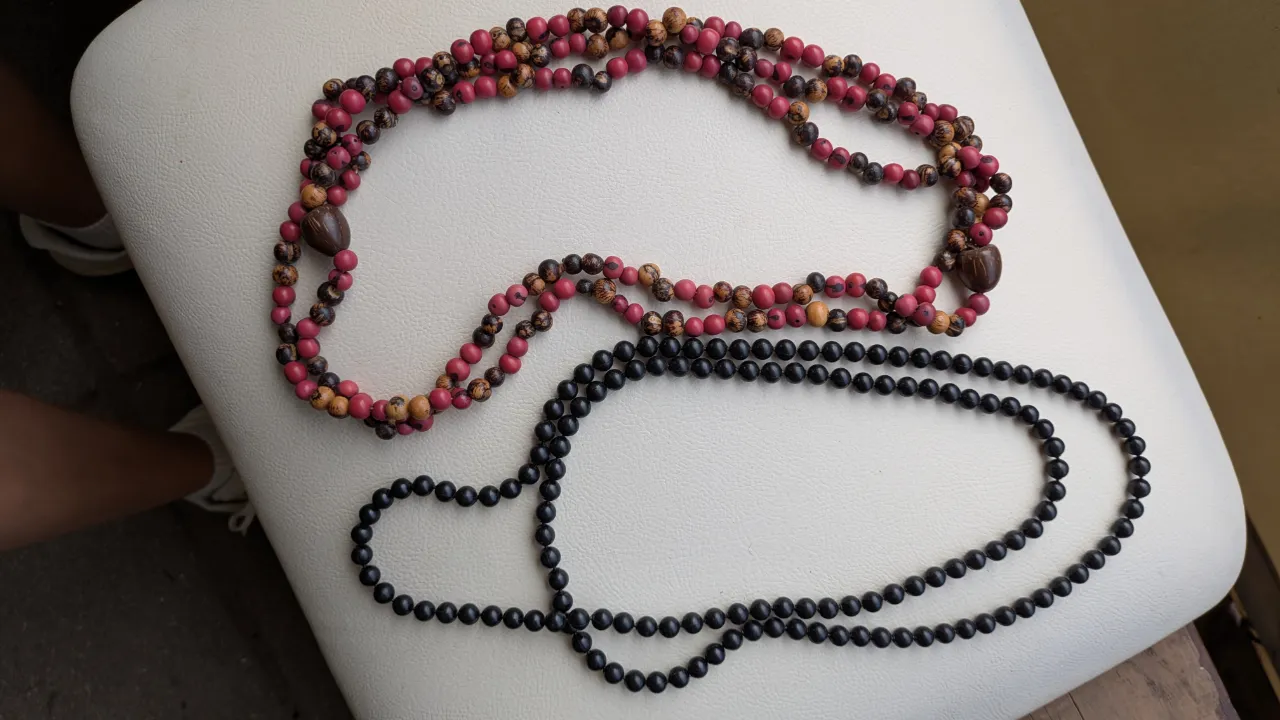 Dos collares de cuentas. Uno es de cuentas pequeñas en tonos fucsia y madera. El otro es de cuentas negras.