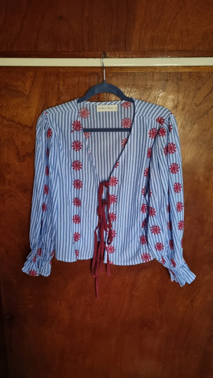 Blusa a rayas celestes y blancas con bordado de flores rojas. Tiene mangas largas con volados y se ata al frente con tiras de tela. Un modelo súper fresco y canchero para usar en cualquier ocasión.