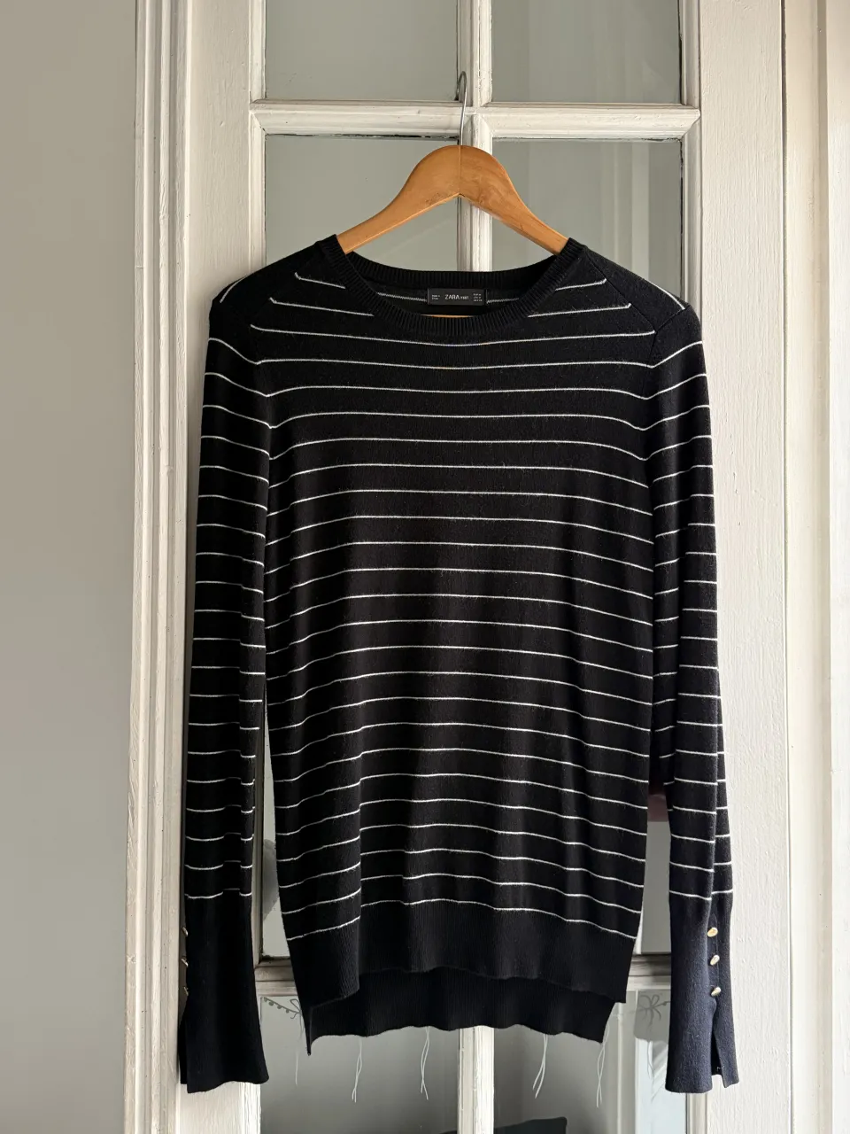 Sweater negro con finas rayas blancas horizontales. Cuello redondo. Puños con tres botones dorados y tajos laterales.