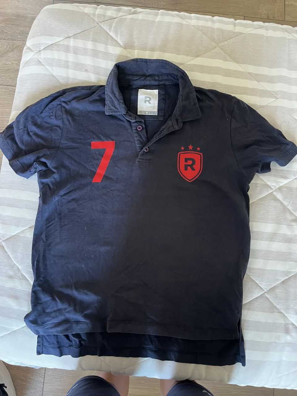 Remera tipo polo de color azul marino con cuello y botones. Tiene un número rojo grande "7" estampado en el frente y un logo rojo con la letra "R" y tres estrellas en el lado superior derecho.