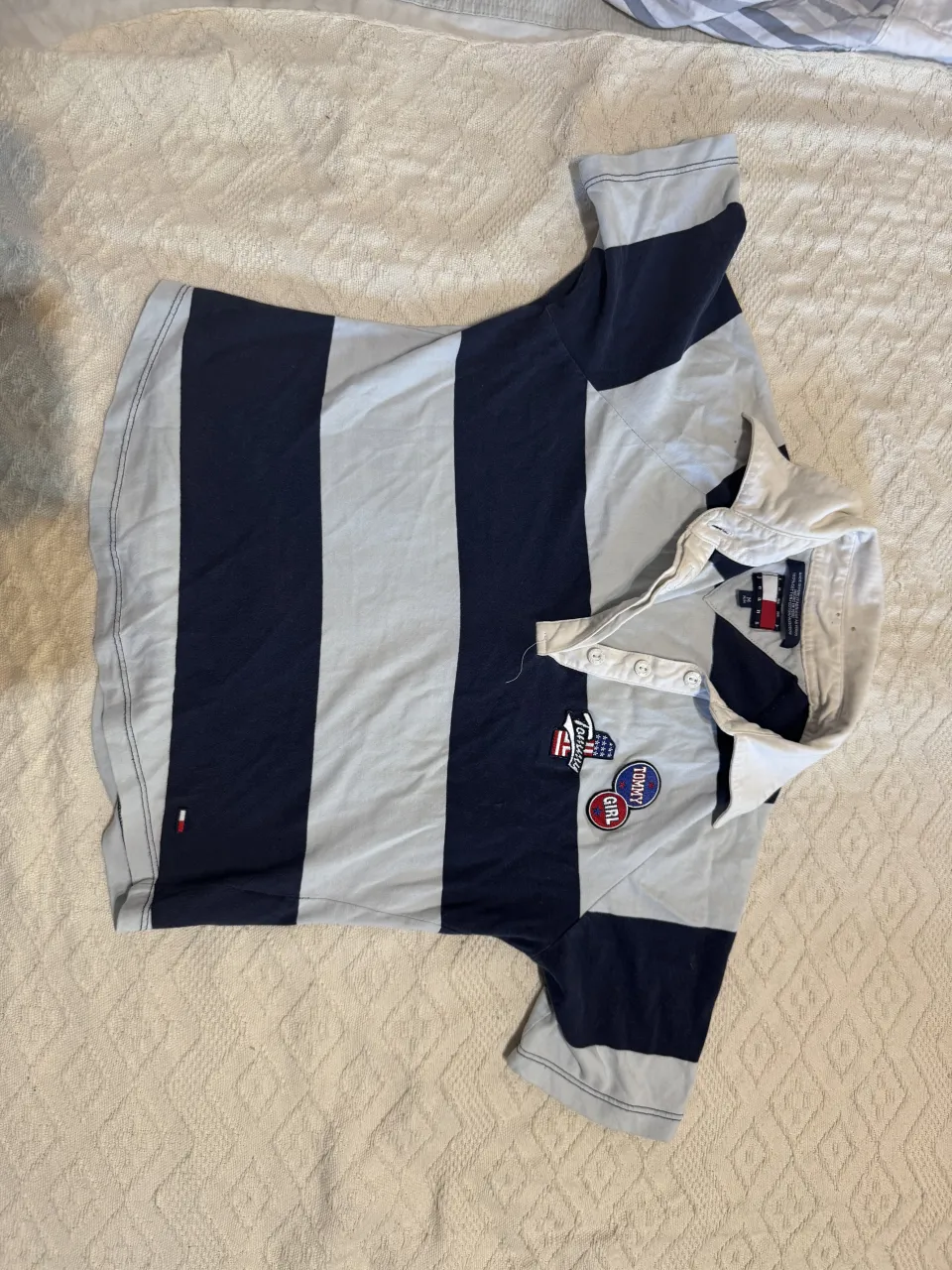 Remera a rayas verticales azul marino y celeste, con cuello blanco y botones. Tiene parches bordados en el frente con el logo de la marca.
Queda entalladita
