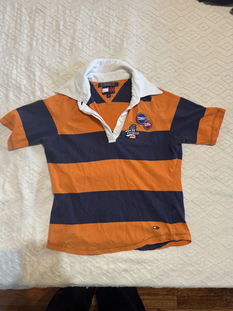 Remera a rayas naranjas y azul marino con cuello blanco y botones. Tiene parches de la marca en el frente.