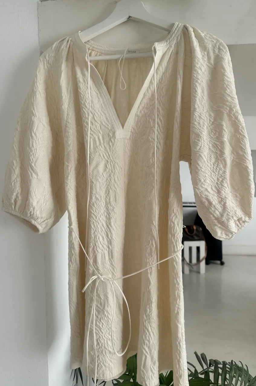 Vestido de tela color crudo con mangas 3/4 abullonadas y escote en V con cordones. Posee un cinturón fino del mismo material.