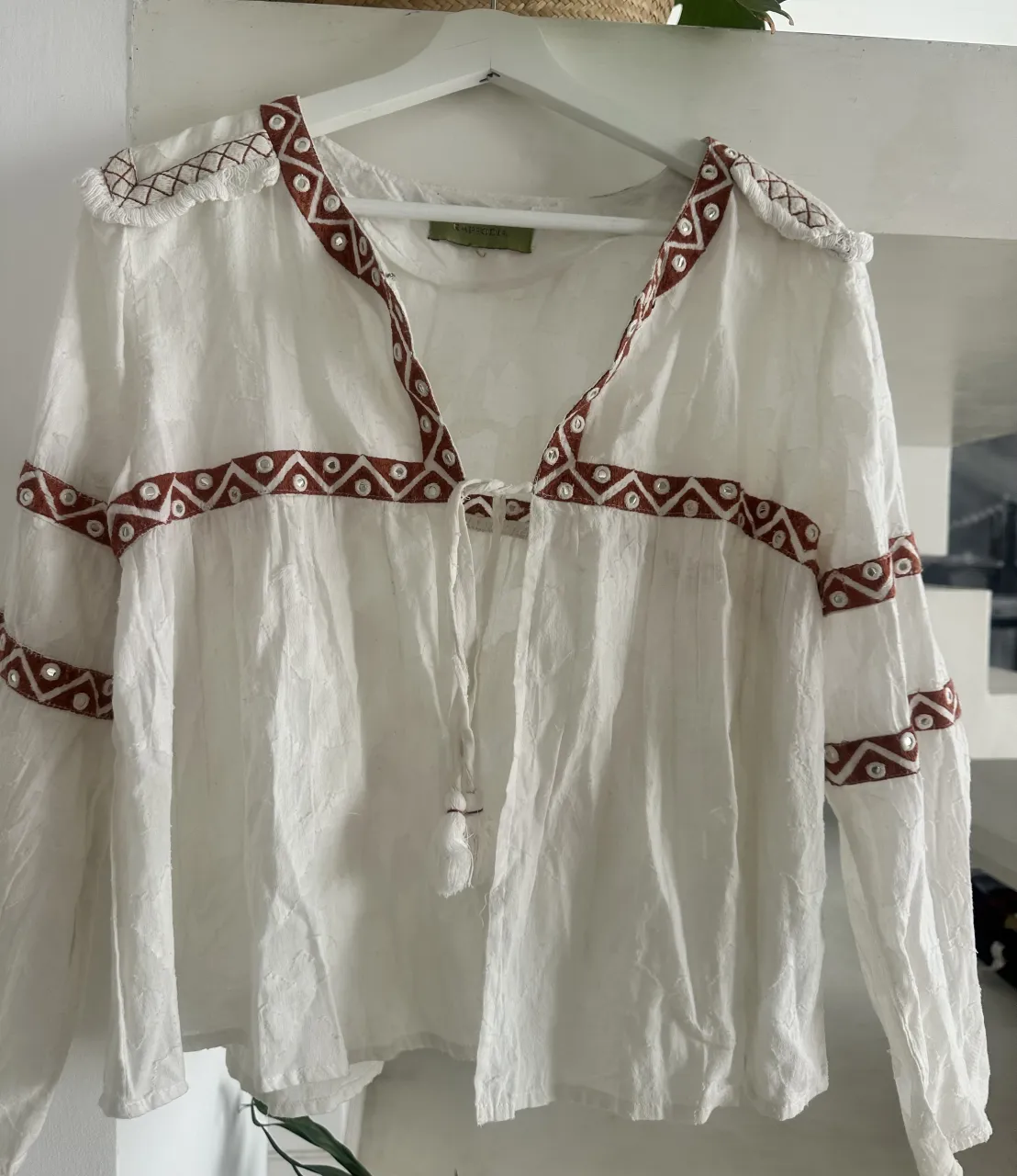 Blusa blanca de manga larga con detalles bordados en color marrón y pequeños espejos incrustados. Posee escote en V con cordones y borla.