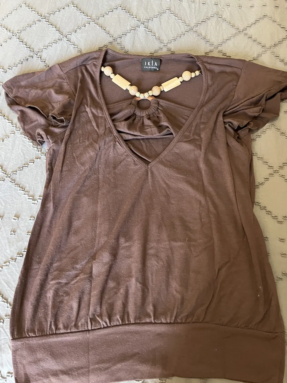 Remera marrón de manga corta con detalles de cuentas de madera en el cuello. Tiene un diseño único con un aro y fruncido en la parte delantera, dándole un toque bohemio y original. Perfecta para un look casual y chic.