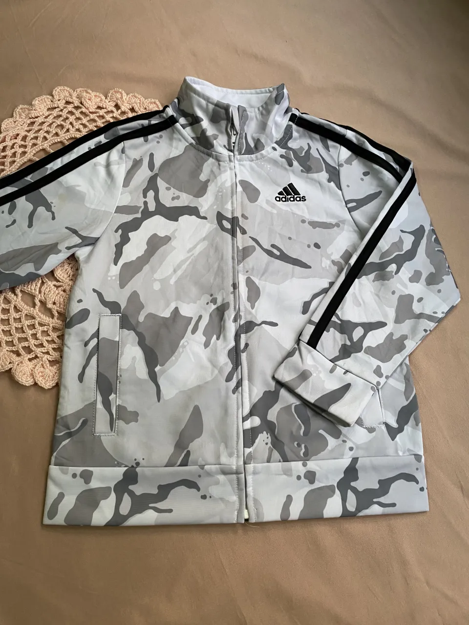 Campera deportiva con cierre y cuello alto. Posee un estampado de camuflaje en tonos grises y blancos, con las tres tiras icónicas de la marca en las mangas. Logo de la marca visible en el frente.