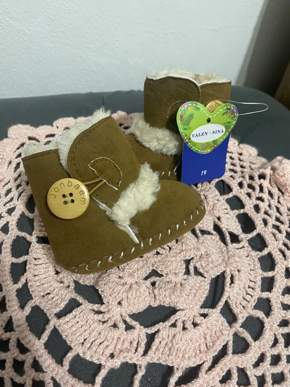 Botitas de bebé de gamuza color marrón con detalles de corderito en la parte superior y un botón de madera con la inscripción 'Jonbaem'.
