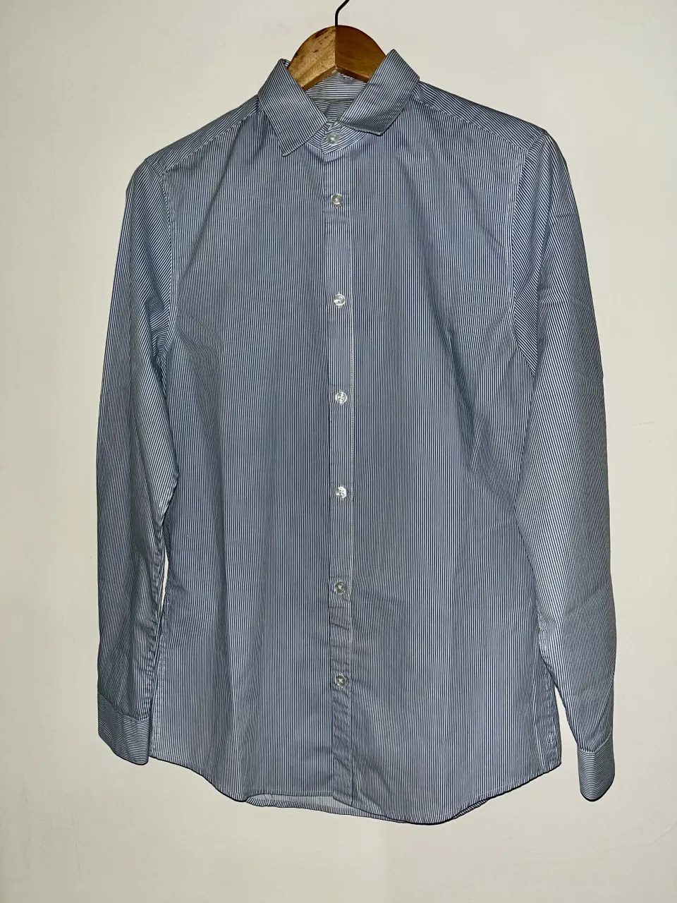 Camisa de manga larga a rayas verticales finas en tonos azul y blanco. Posee botones al frente y en los puños.