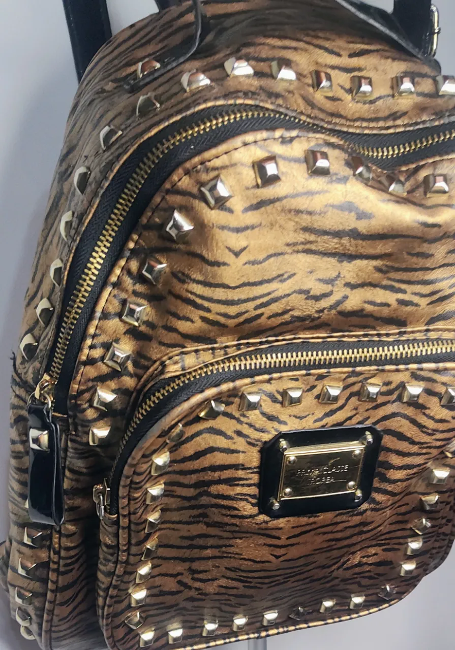 🎒  Mochila 🎒 de la línea Prima Classe de Tropea, caracterizada por su diseño en animal print de cebra (o "acebrado") en tonos ocre y negro, decorada con tachas doradas cuadradas.  con estampa de tigre y tachas metálicas cuadradas. Posee un bolsillo frontal con cierre y un bolsillo principal con cierre.